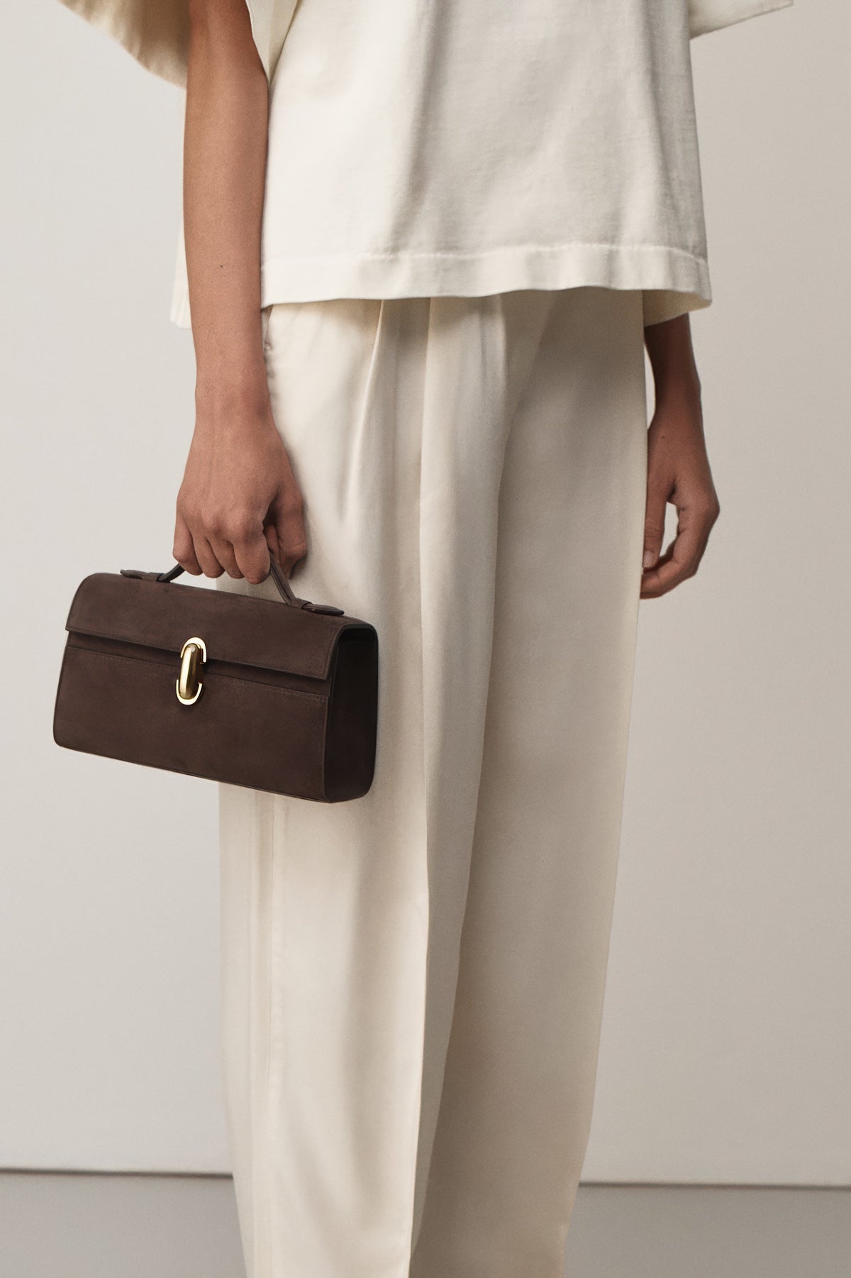 on model view of a coffee brown suede top handle bag with a gold clasp on a white background