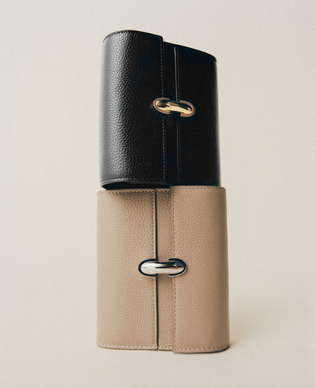 editorial image of a black wallet with a gold clasp on top of a bone wallet with silver clasp on a white background