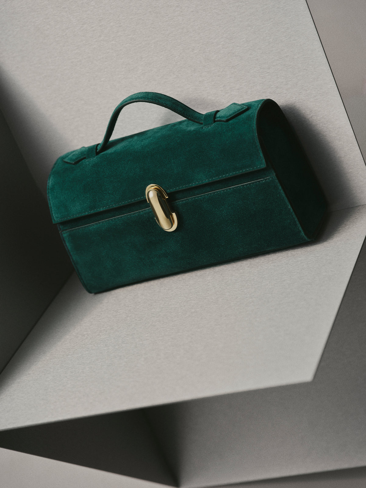 campaign image of pine green suede bag with gold clasp on a grey background