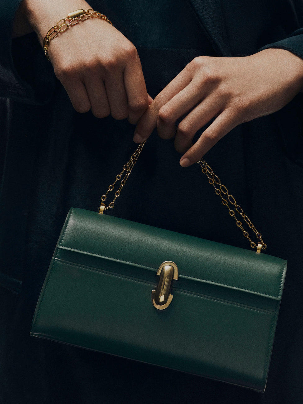 campaign image of model holding pine green leather bag with gold clasp and gold chain on a black background